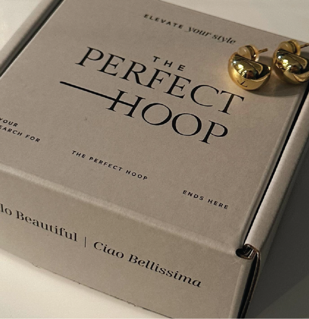 A slightly tilted cubic package with the title 'The Perfect Hoop' highlighted is positioned in the center of the image. The package is desaturated beige in color. In the upper right corner of the image, two branded earrings are displayed. The background color is light beige, resembling almond milk. Additional information about the product and brand is placed at the top and bottom of the package.