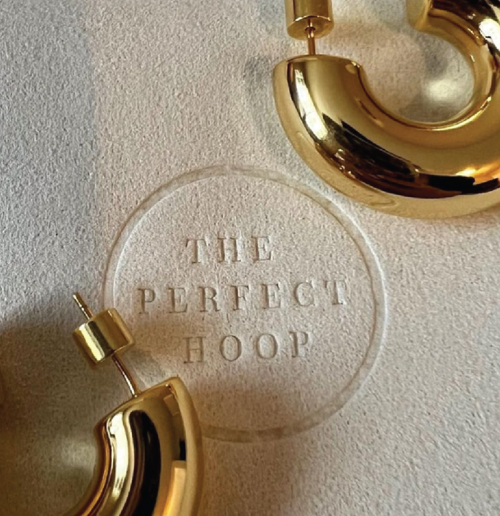 In the centered background, there is a paper with the brand 'The Perfect Hoop' marked with a circumscribed textured print, created using the hot stamping technique. Partially visible on the front of the paper, in the upper right and lower left corners, are two golden rings produced by the brand.