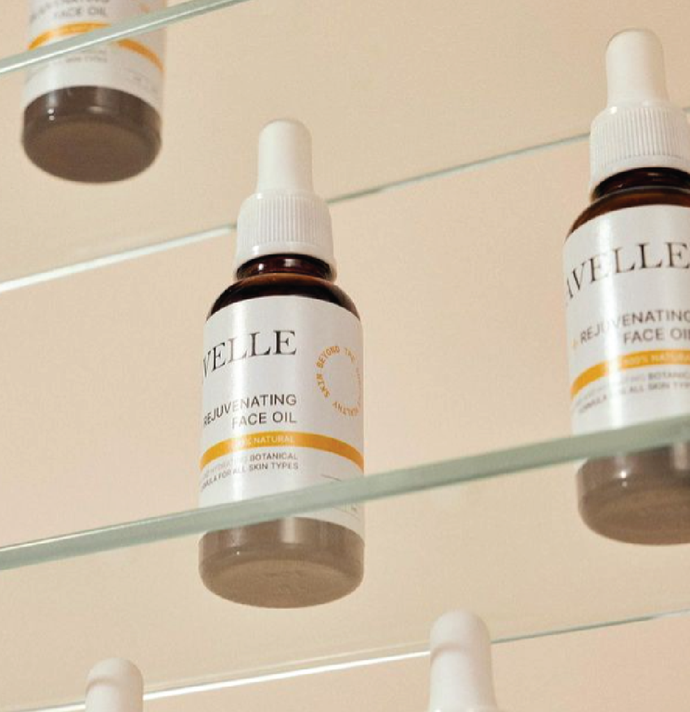 On a glass shelf, there is a slightly inferior view of three dark glass bottles with cloud-white dropper caps. The bottles belong to the 'Avelle' brand facial serum. The background of the image is light beige in color.